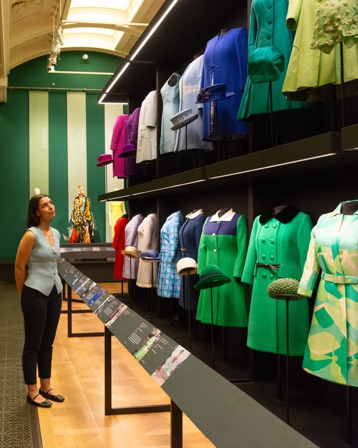 A person gazes at an exhibition of vibrant vintage clothing displayed on mannequins, including green, blue, and purple outfits. The display is in a well-lit room with green walls, showcasing a variety of textures and styles. In the background, there is a figure wearing an intricately patterned outfit.